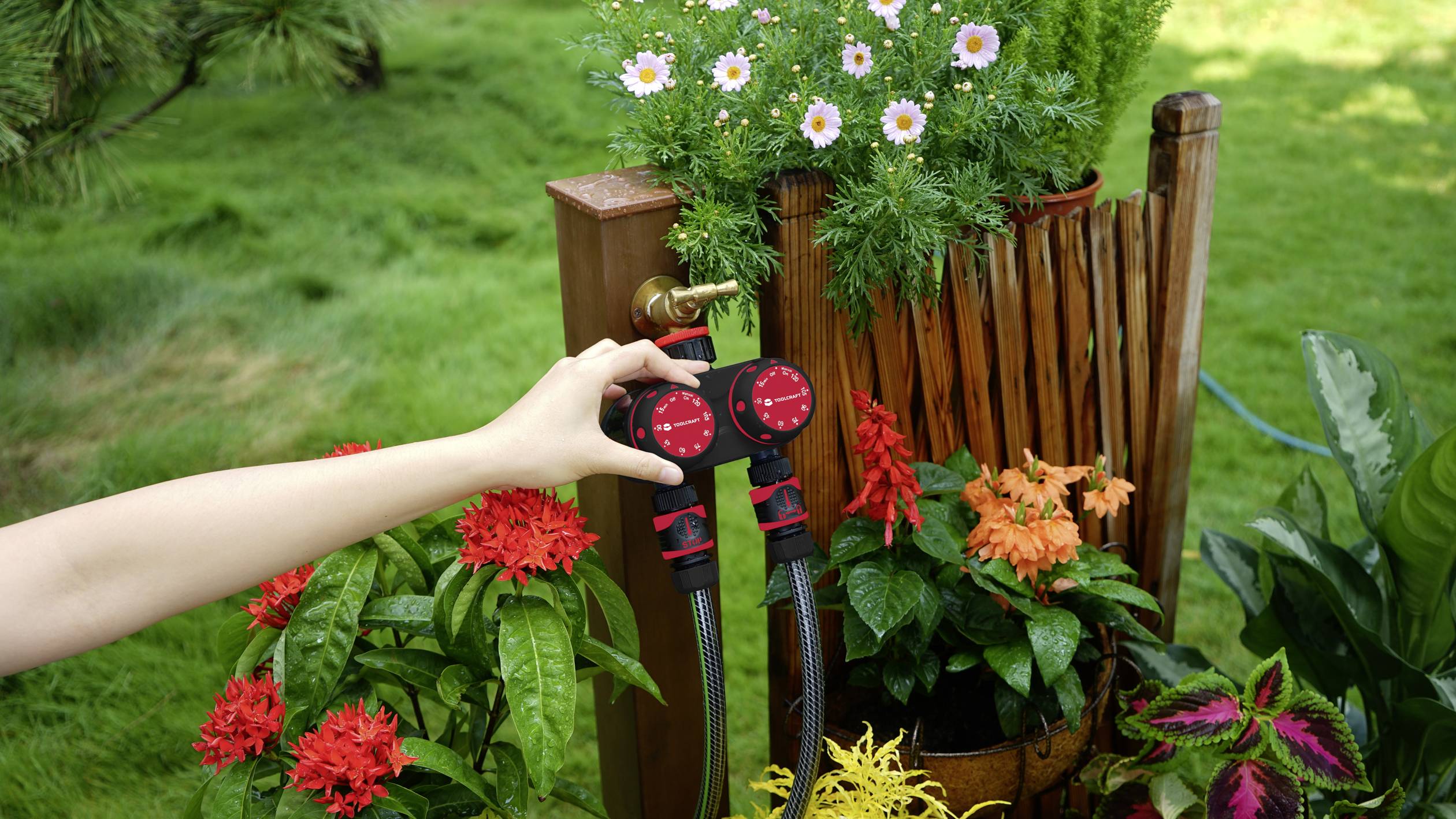 A person is adjusting two red water valves on an outdoor tap, surrounded by flowering plants and a green lawn in the background.
