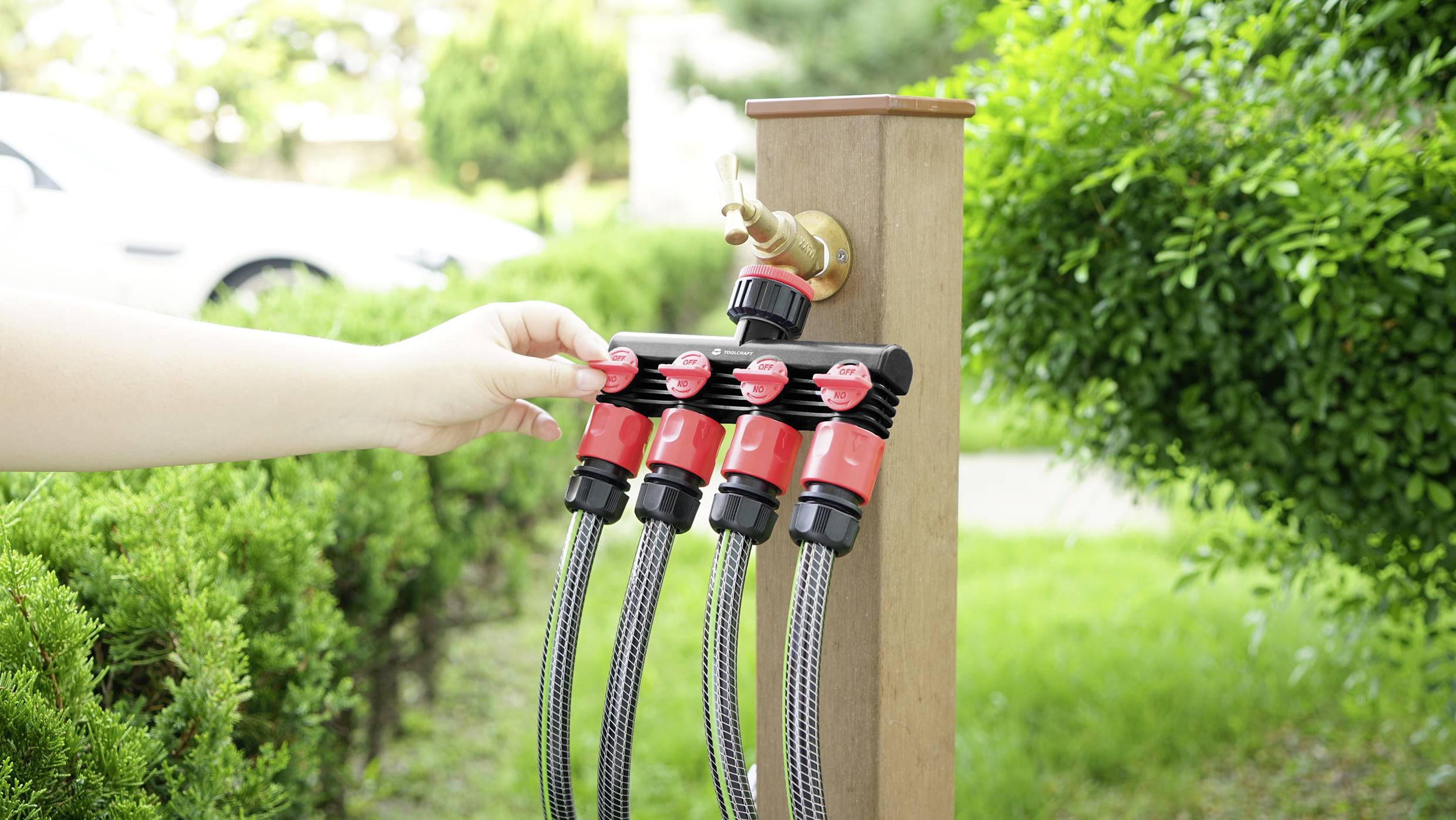 A hand turns the water tap valve with five hoses in the greenery.