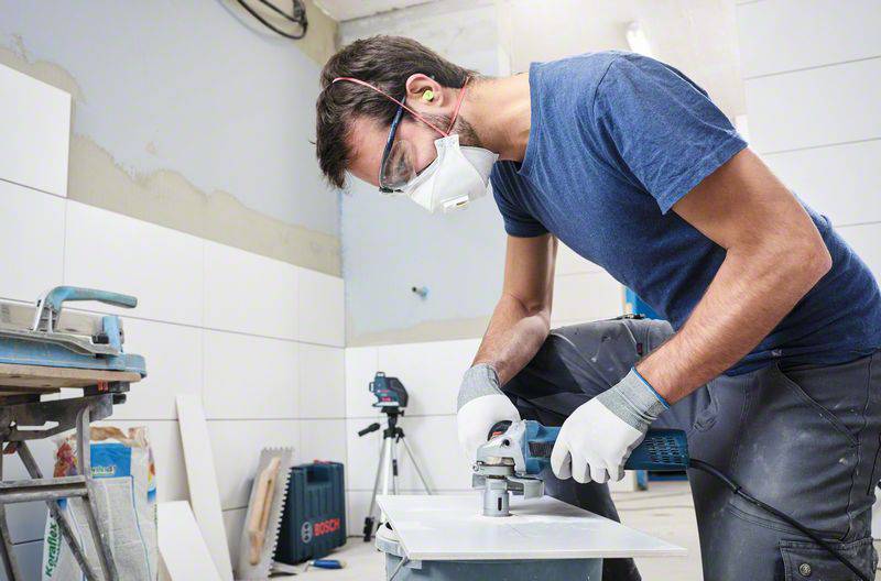A tradesman wearing safety glasses and a mask is cutting a tile with an electric saw in a workshop environment.