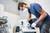 A tradesman wearing safety glasses and a mask is cutting a tile with an electric saw in a workshop environment.