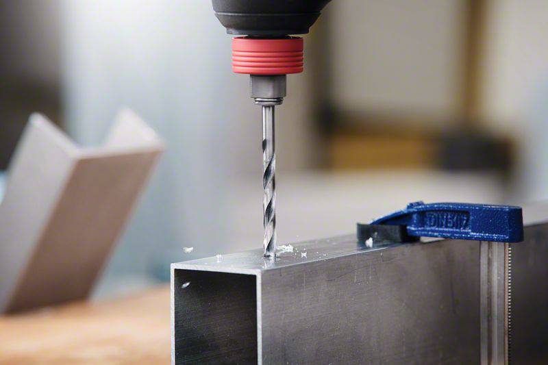 A drill is boring a hole into a metal square. Another piece of metal is blurred in the background to the side.