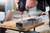 A hand is holding a Bosch cordless drill, drilling a hole into a metal profile that is clamped to a wooden table.