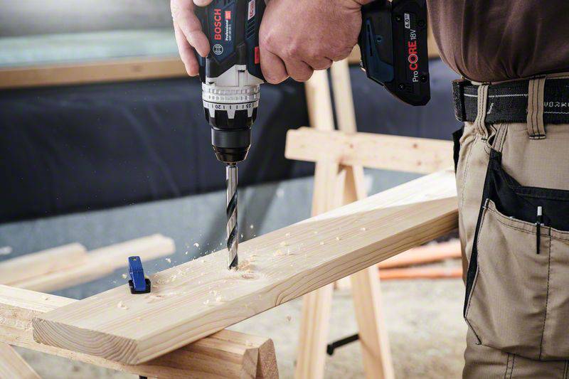 A person is drilling a hole into a wooden board with a Bosch drill, which is placed on a workbench and surrounded by wood shavings.