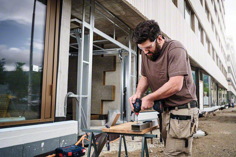 A construction worker in a brown T-shirt and work trousers is using a power drill to secure metal profiles on a building site. A building is visible in the background.