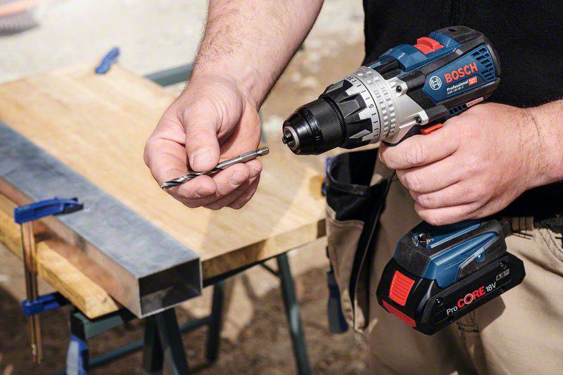 A person is holding an electric drill and a drill bit. In the background, a vice with wood and metal can be seen.
