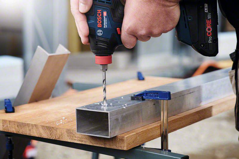 A person is drilling a hole in a rectangular metal tube using a Bosch cordless drill driver on a wooden table.