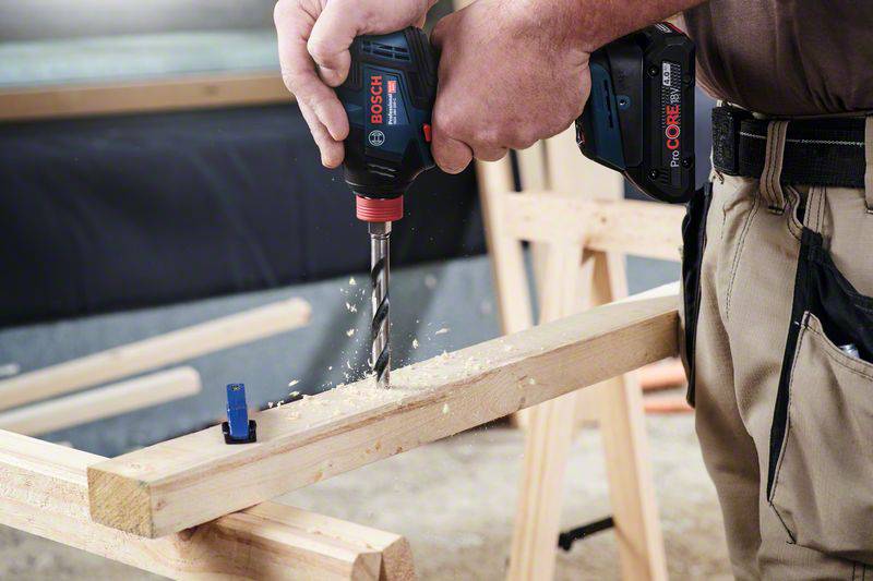 A tradesman is drilling a hole in a wooden board on a construction site. Wood shavings fall as the drill works.