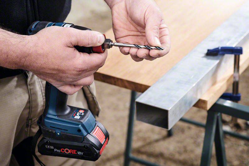 A person is holding a drill and inserting a drill bit. A piece of metal lies on a table, secured with a clamp.
