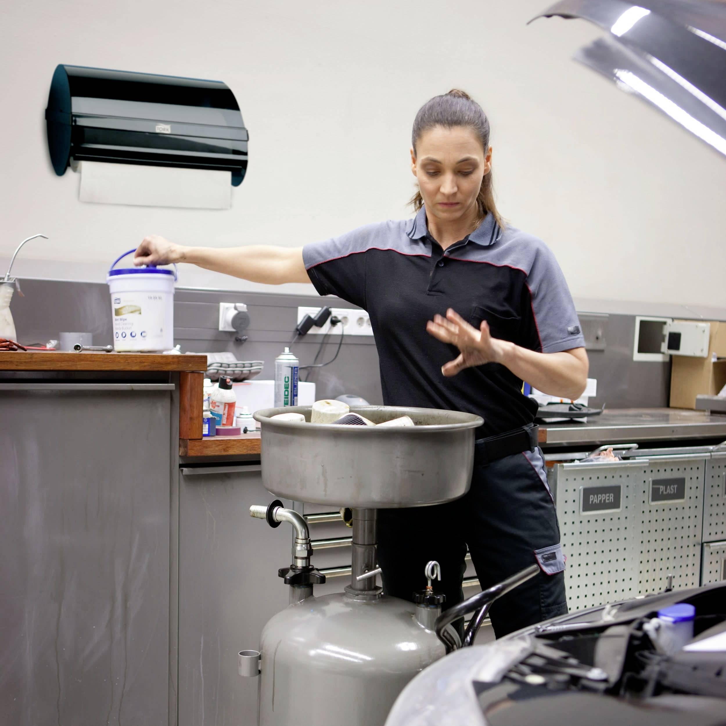 Woman in workshop, holding paint bucket next to compressed air tank, likely preparing for painting; air conditioning unit is present in the room.
