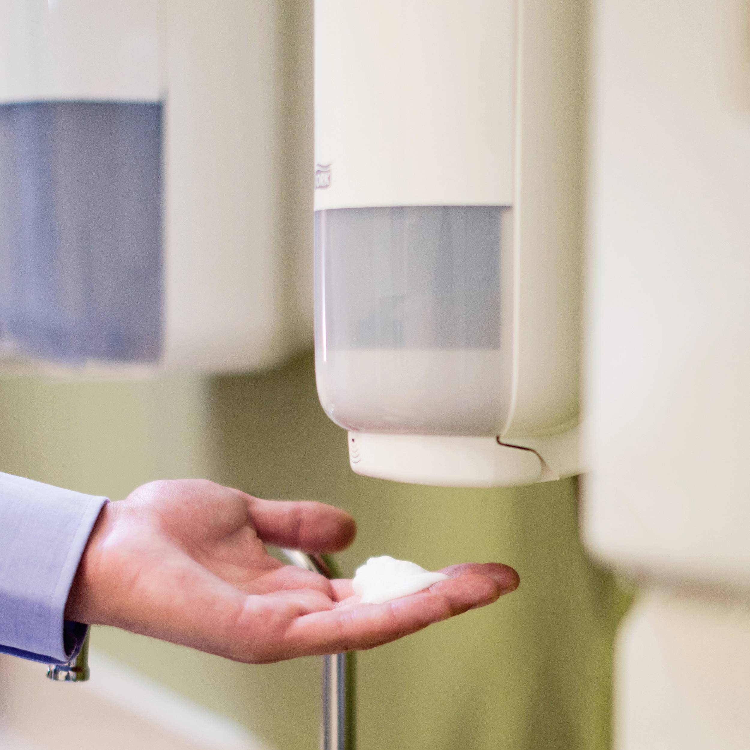 A person presses the soap dispenser on the wall and pumps liquid soap into their hand in a public washroom.