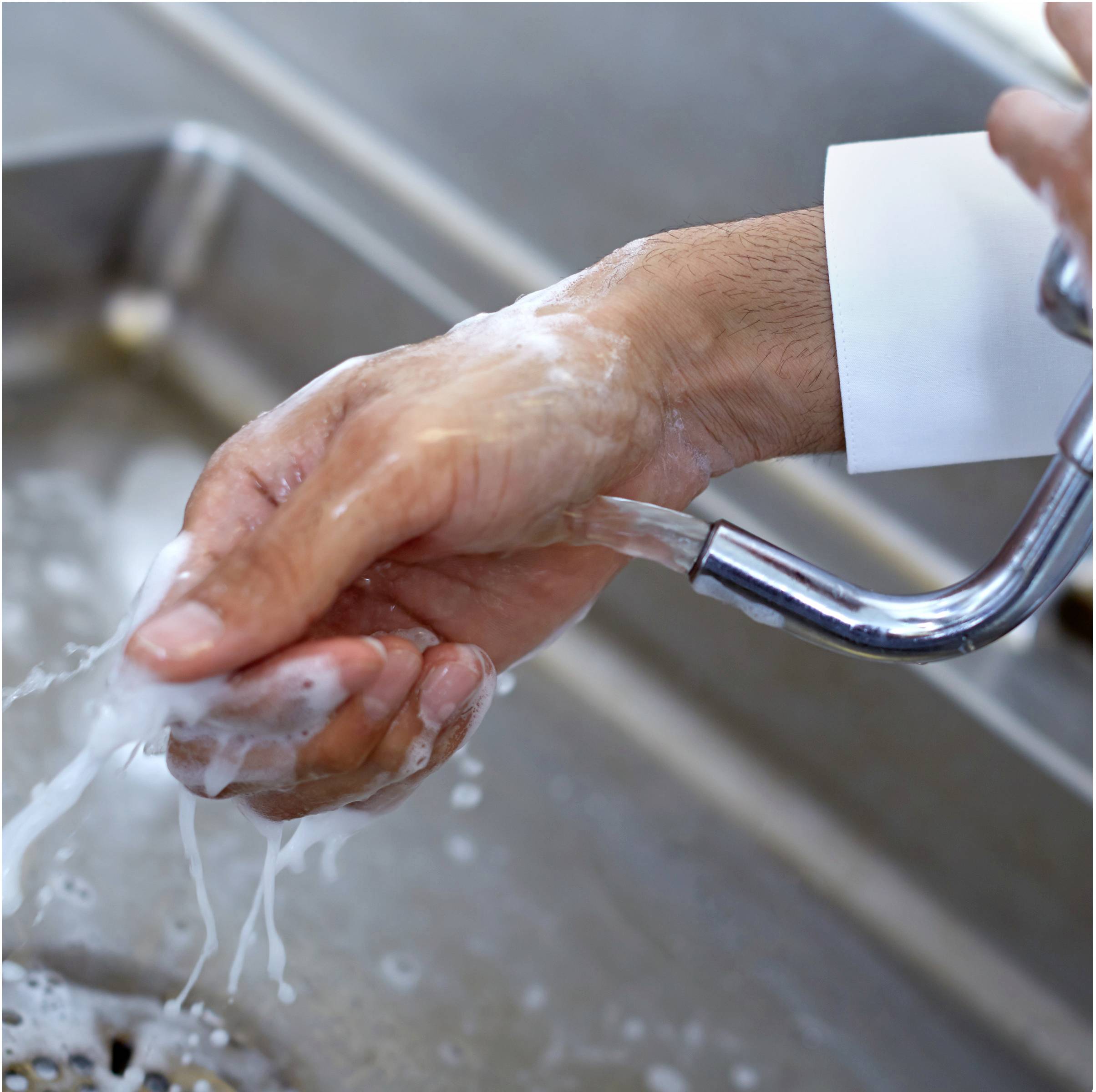 A person is washing their hands under running water in a stainless steel sink. Hands are lathered with soap.