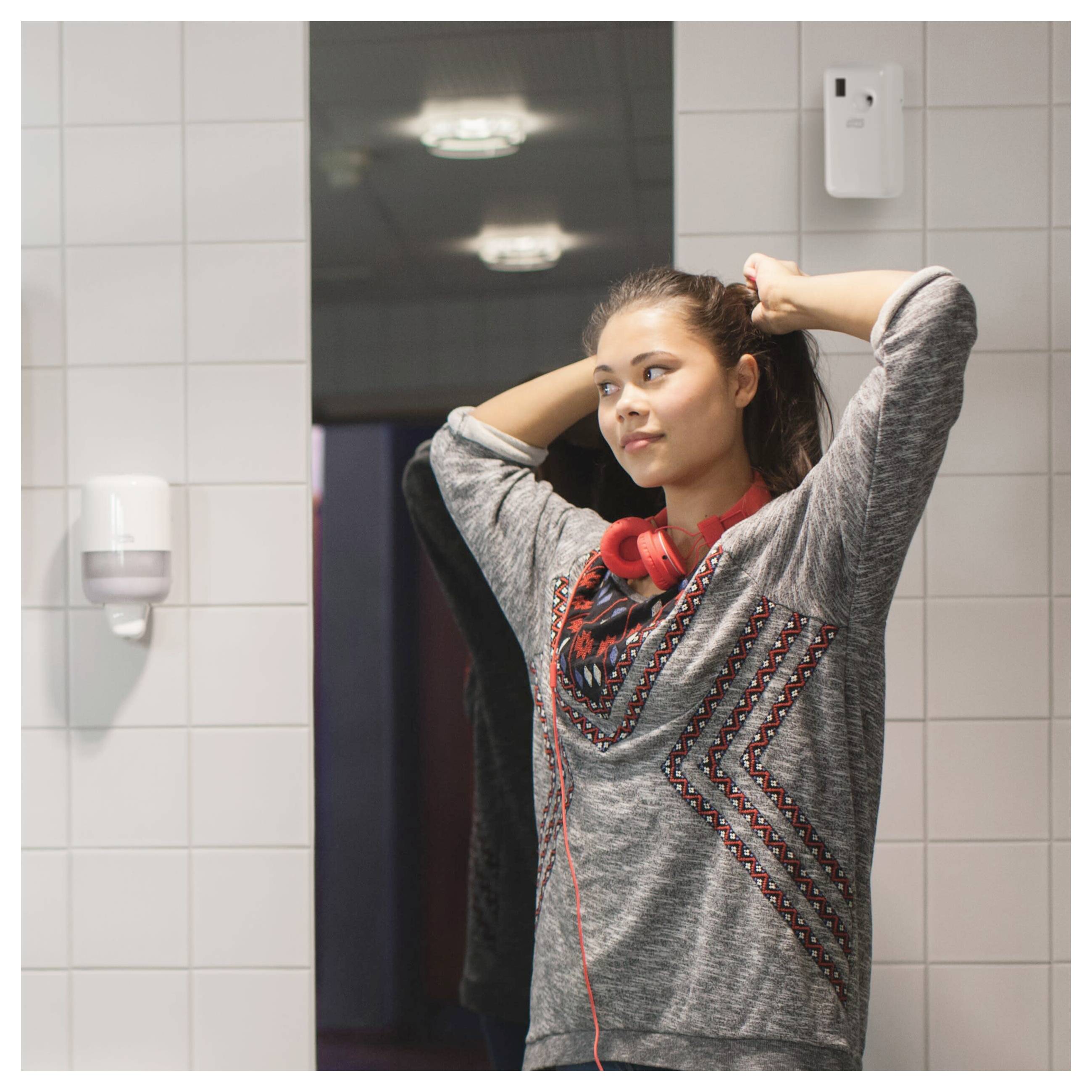 Young woman in a tiled restroom, tying back her hair while wearing red headphones around her neck.