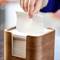 A hand pulls a paper towel from a wooden dispenser on a table.