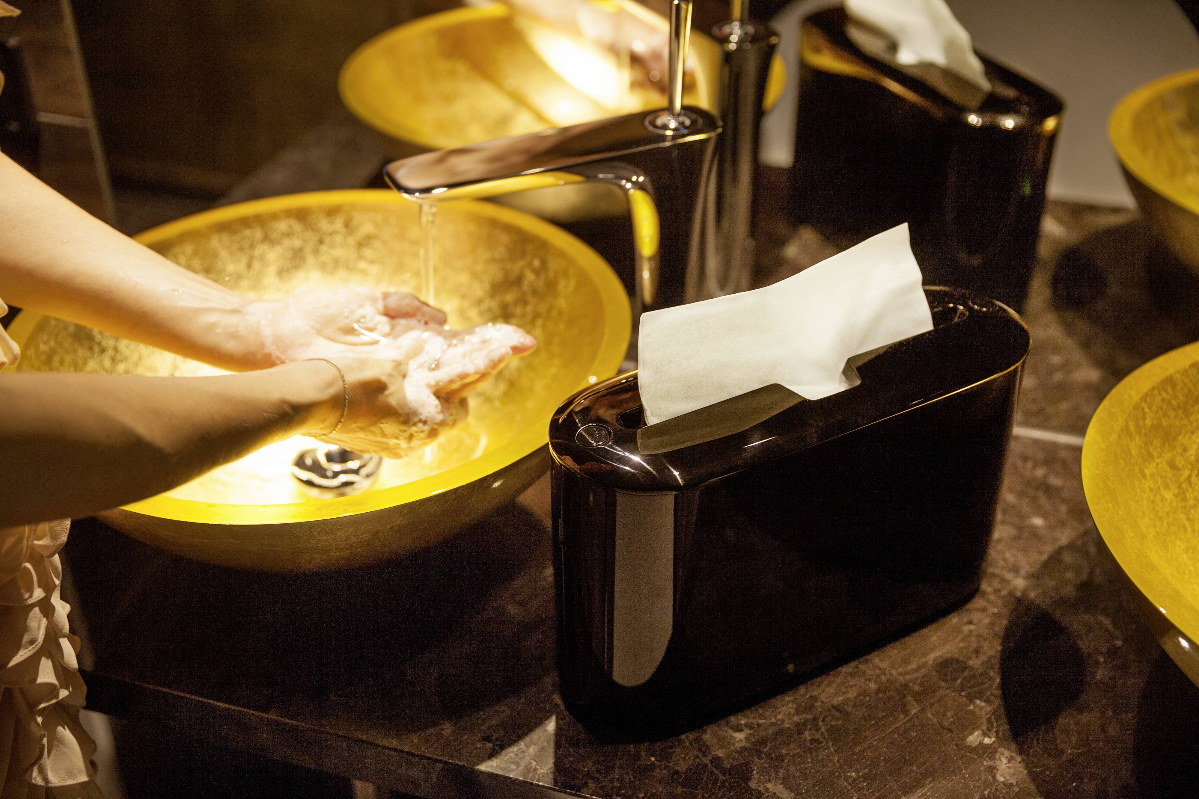 A person is washing their hands under running water in a golden sink; a black dispenser with paper towels is beside it.