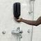 A person is pressing a black soap dispenser on a tiled bathroom wall, near a shower with metal fittings.