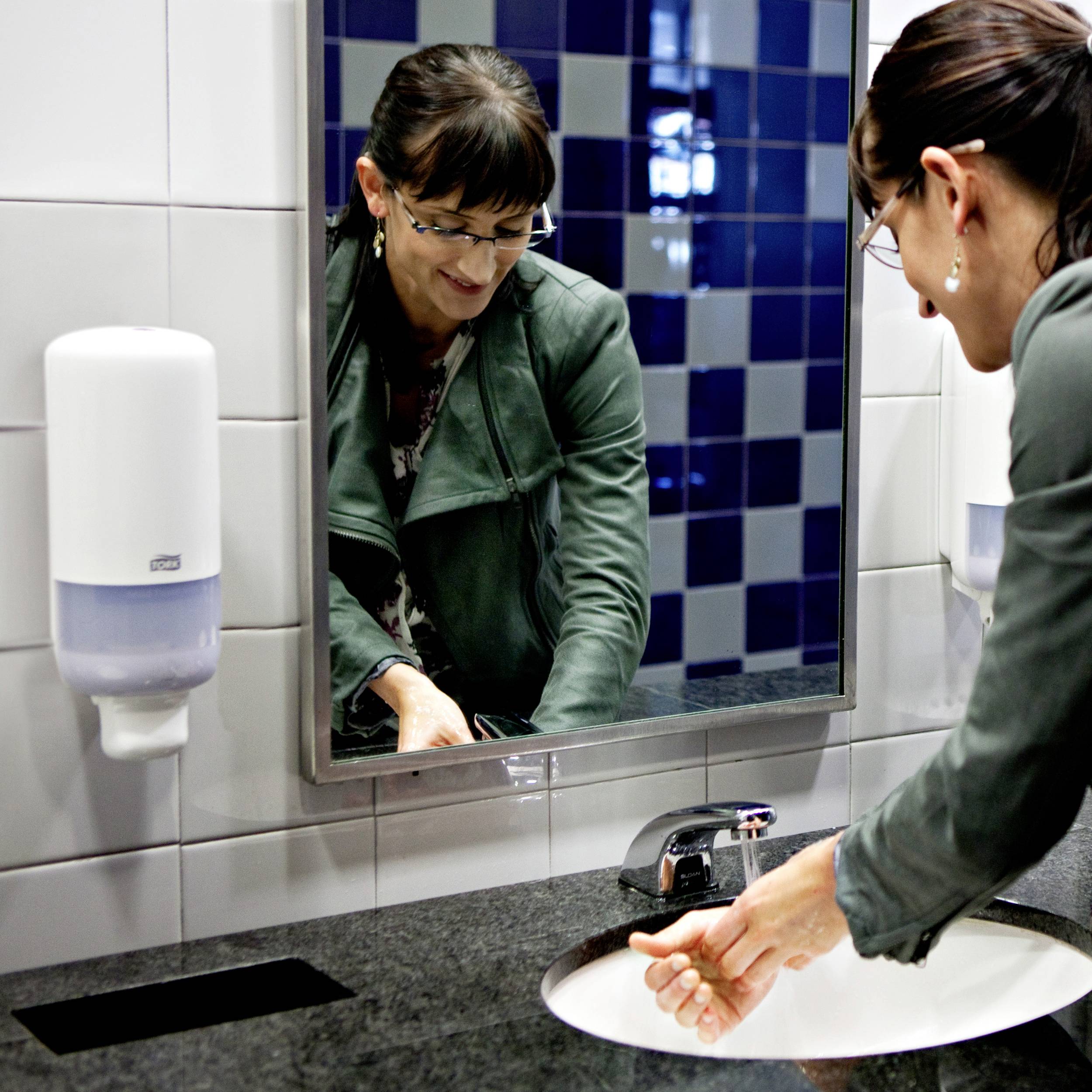 A woman is washing her hands at a sink in a bathroom with a soap dispenser on the wall. She looks into the mirror with a smile.