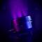 A black lighting device emits violet light. In the background, additional lights are visible, illuminating a stage.