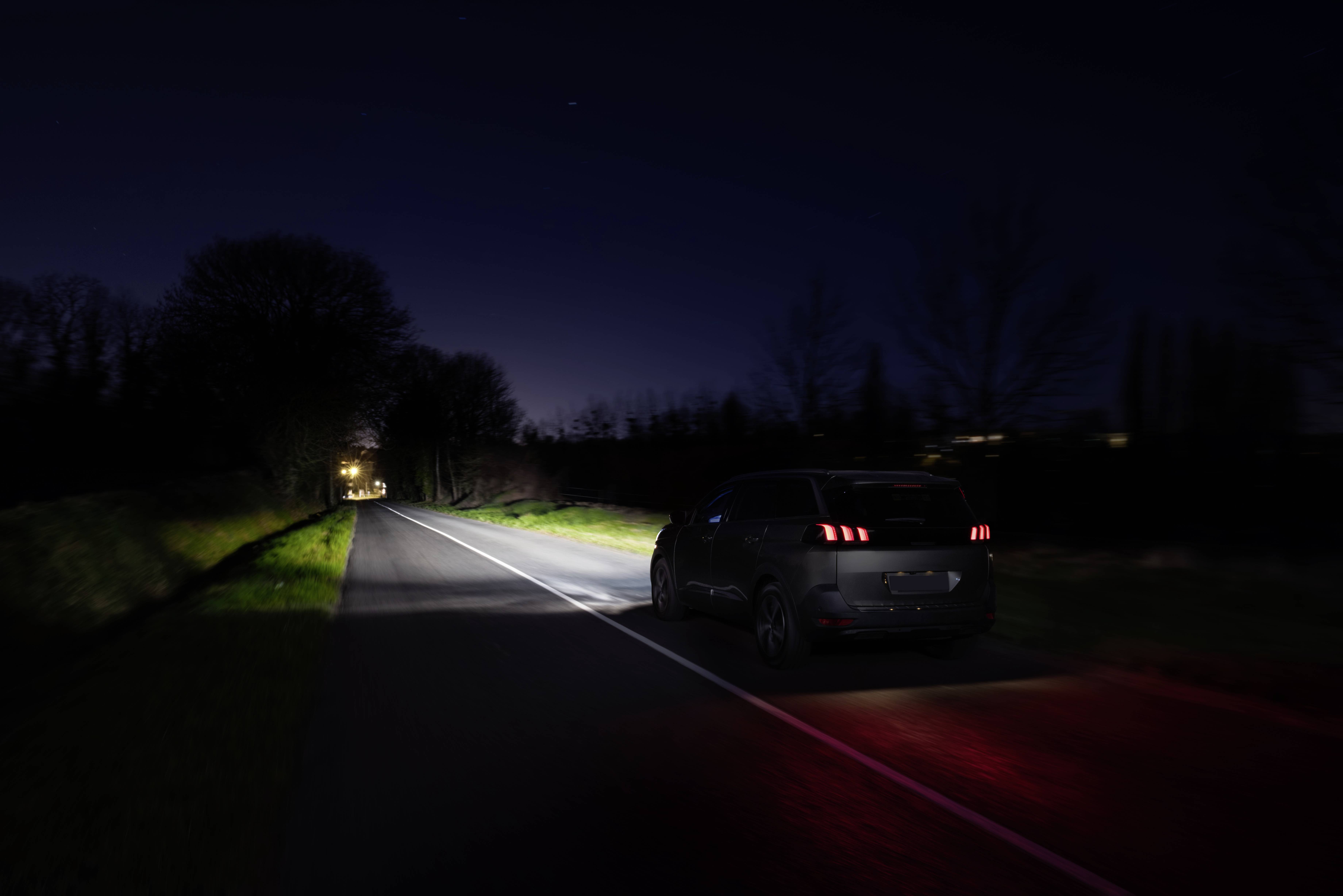 A car is driving along a country road at night. Its rear lights glow red, while the headlamps illuminate the road ahead.