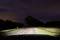 Street at night illuminated by car headlights, leading into a curve with trees and meadows on the sides. Dark, cloudy sky.