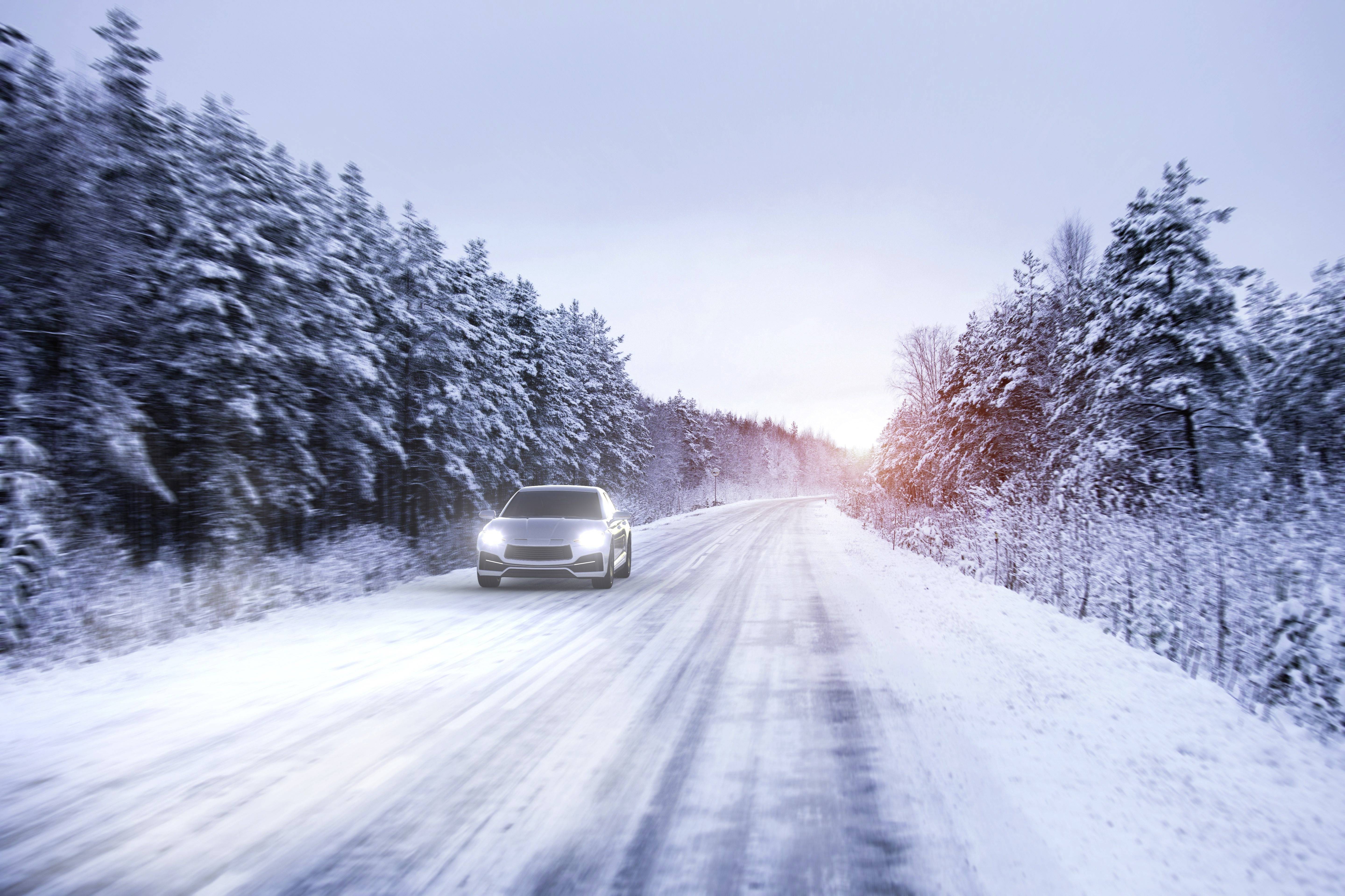 A car is driving on a snow-covered road through a snow-laden forest at sunset. Its headlights are switched on.