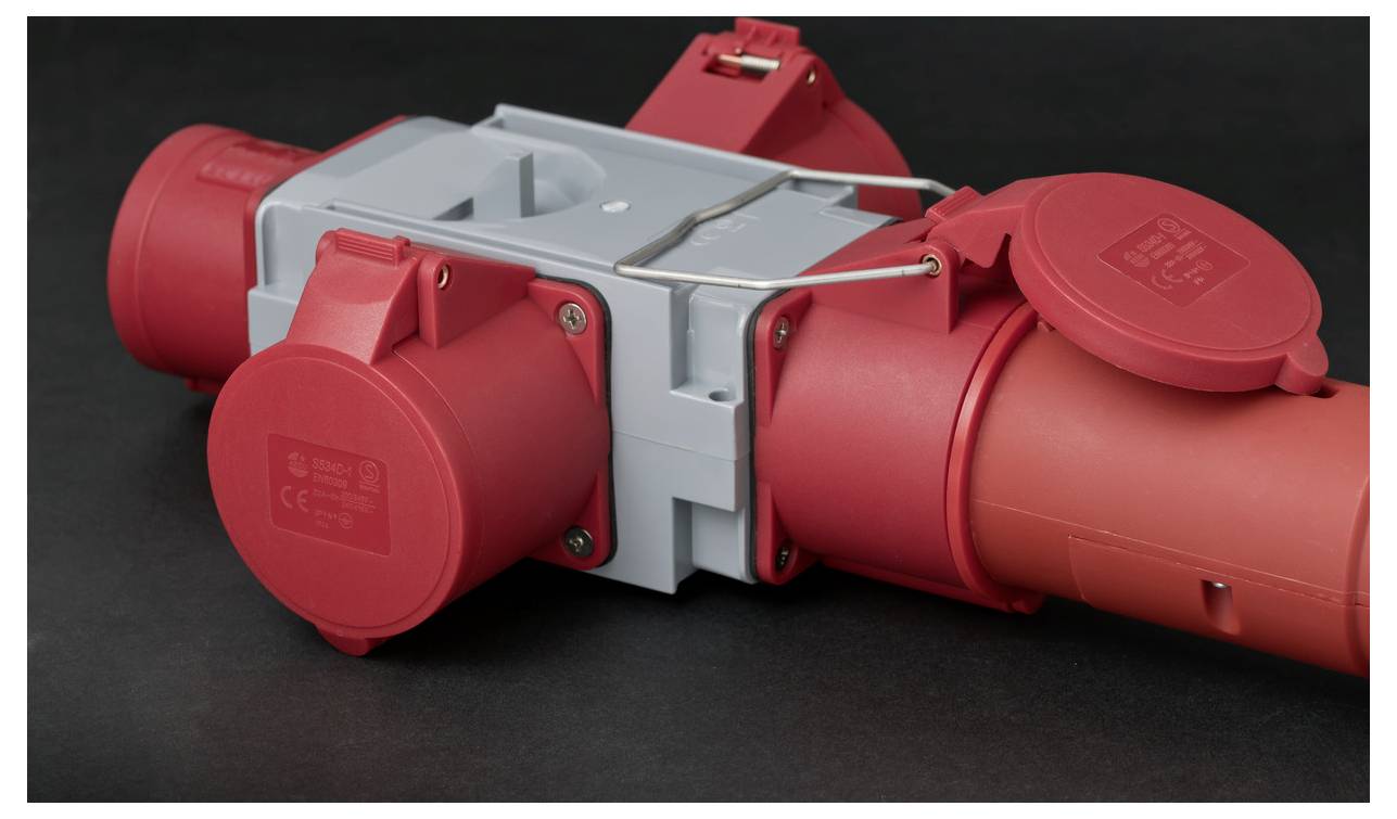 Red industrial electrical power connector with multiple covered outlets and a gray central housing on a dark background.