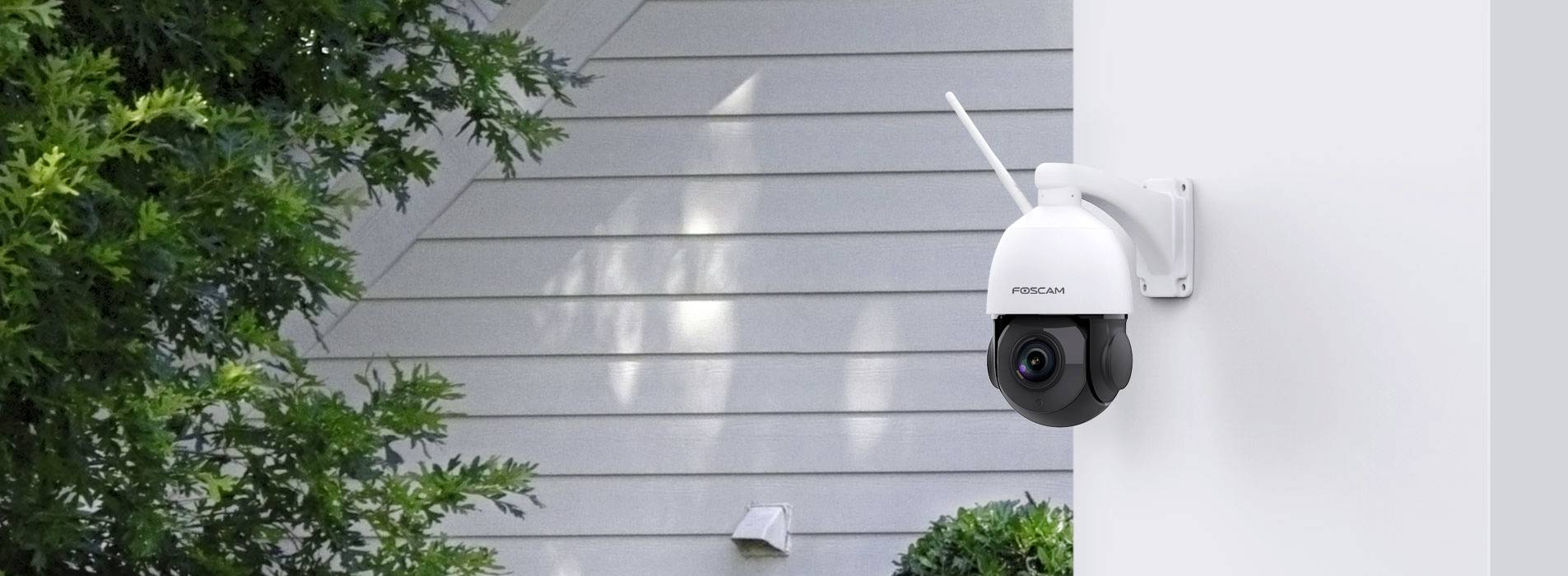 Outdoor camera mounted on a white wall, monitoring the property. Green plants are visible to the left, with another building in the background.