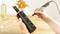 A person is engraving a pattern onto a dark glass bottle using a tool. In the background, wine is placed on a table with flowers.
