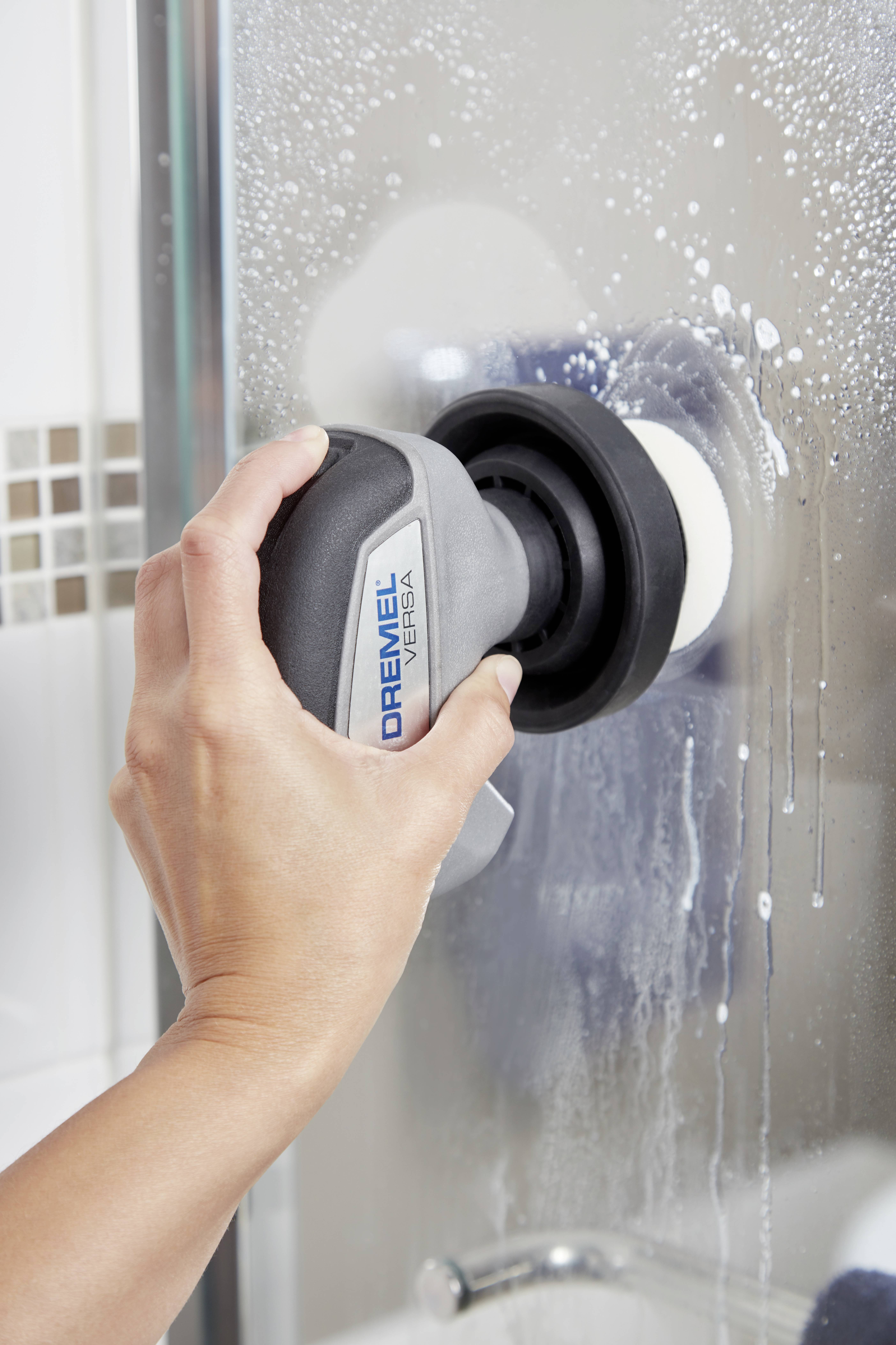 A hand is holding a cleaning device with the label 'DREMEL VERTO', which is being used on a glass shower screen.
