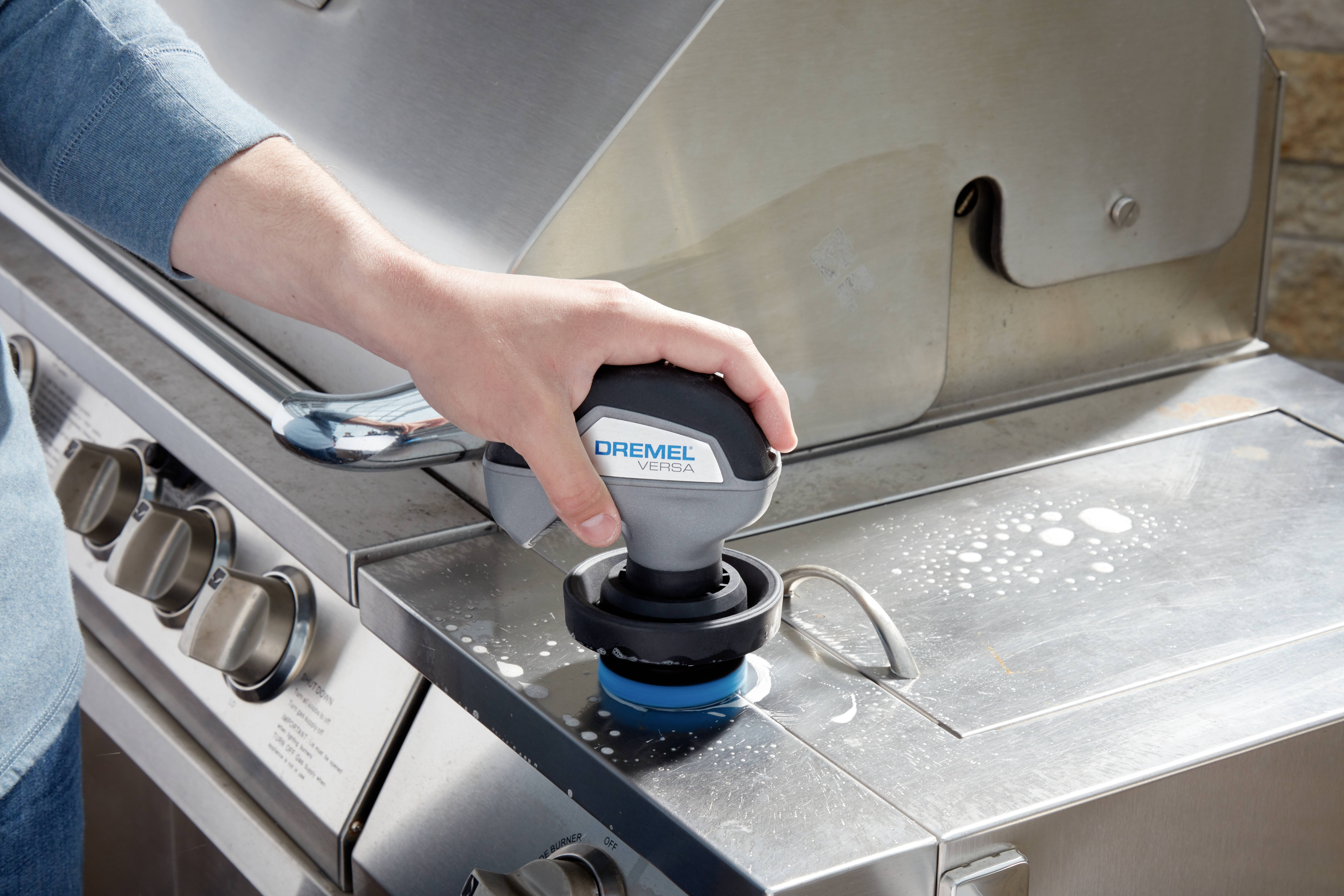 A person is cleaning a stainless steel surface of a barbecue with a Dremel power tool. Foam is visible on the surface.