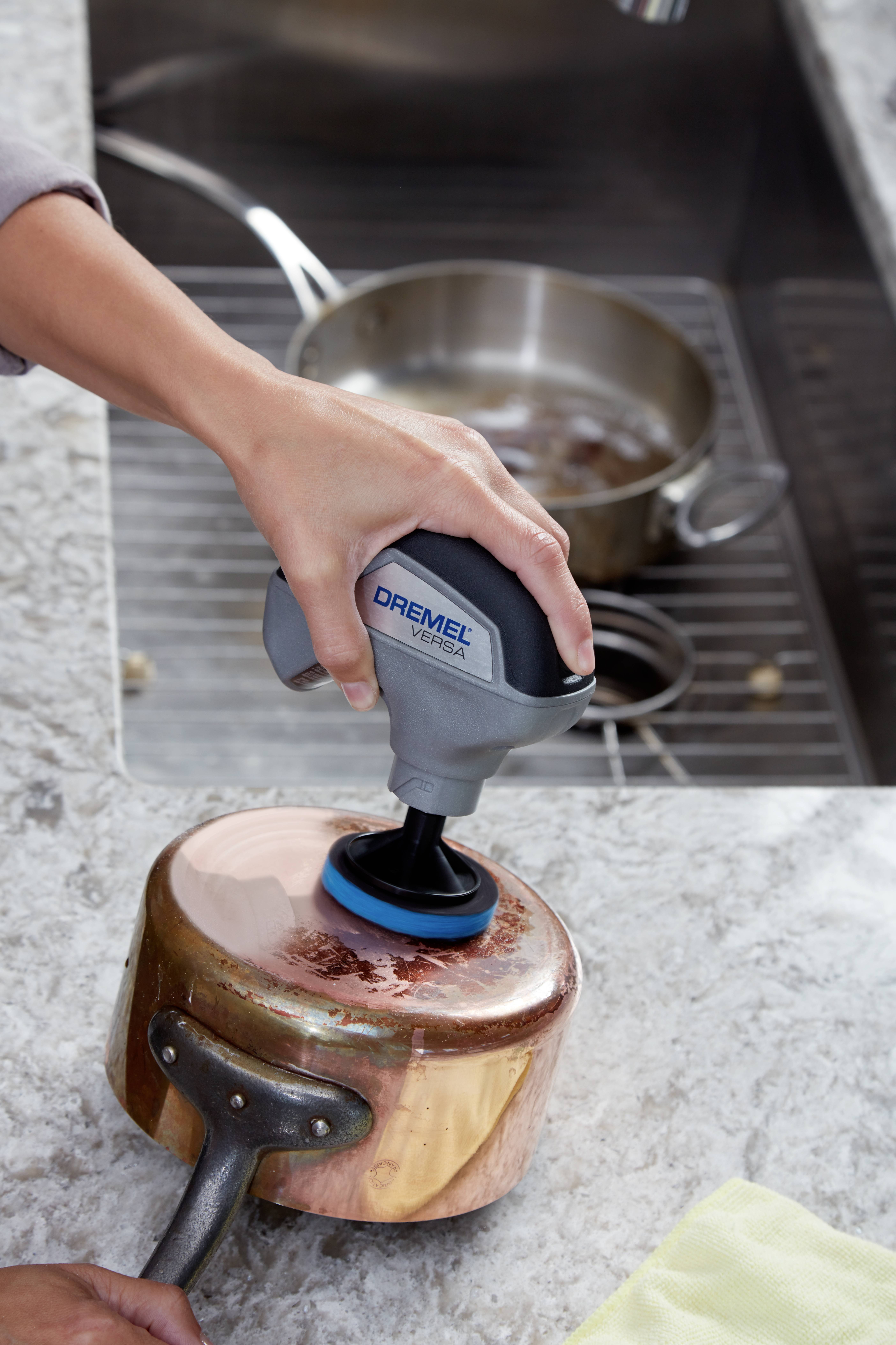 A person is using an electric cleaning device to polish a copper saucepan on a kitchen worktop. In the background, a pot is standing in the sink.