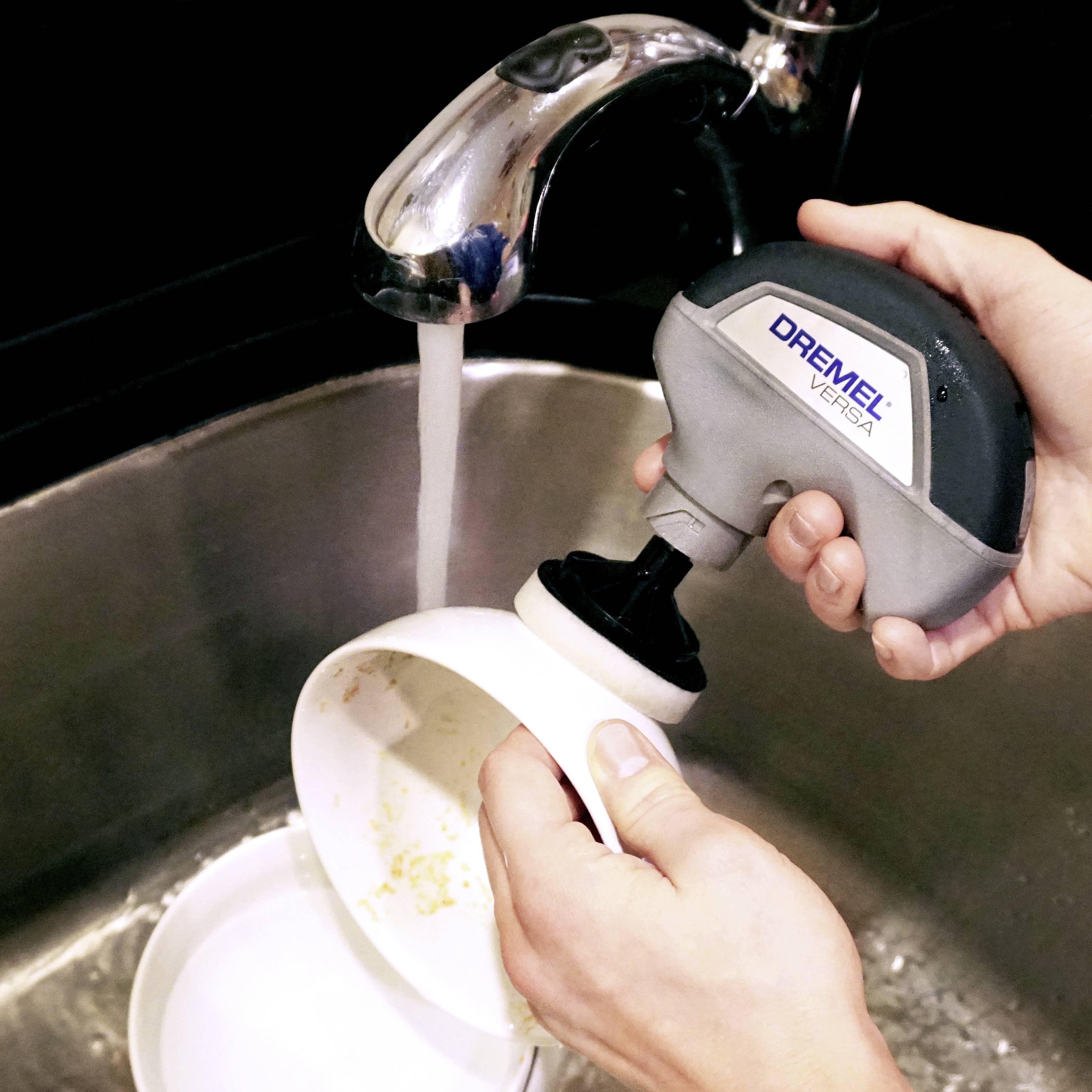 A person is cleaning a cup with an electric cleaning device under running water, whilst a plate is being washed beside it.