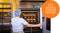 A female baker in work attire opens an oven where several trays of bread rolls are being baked. An icon for baking appears on the right.