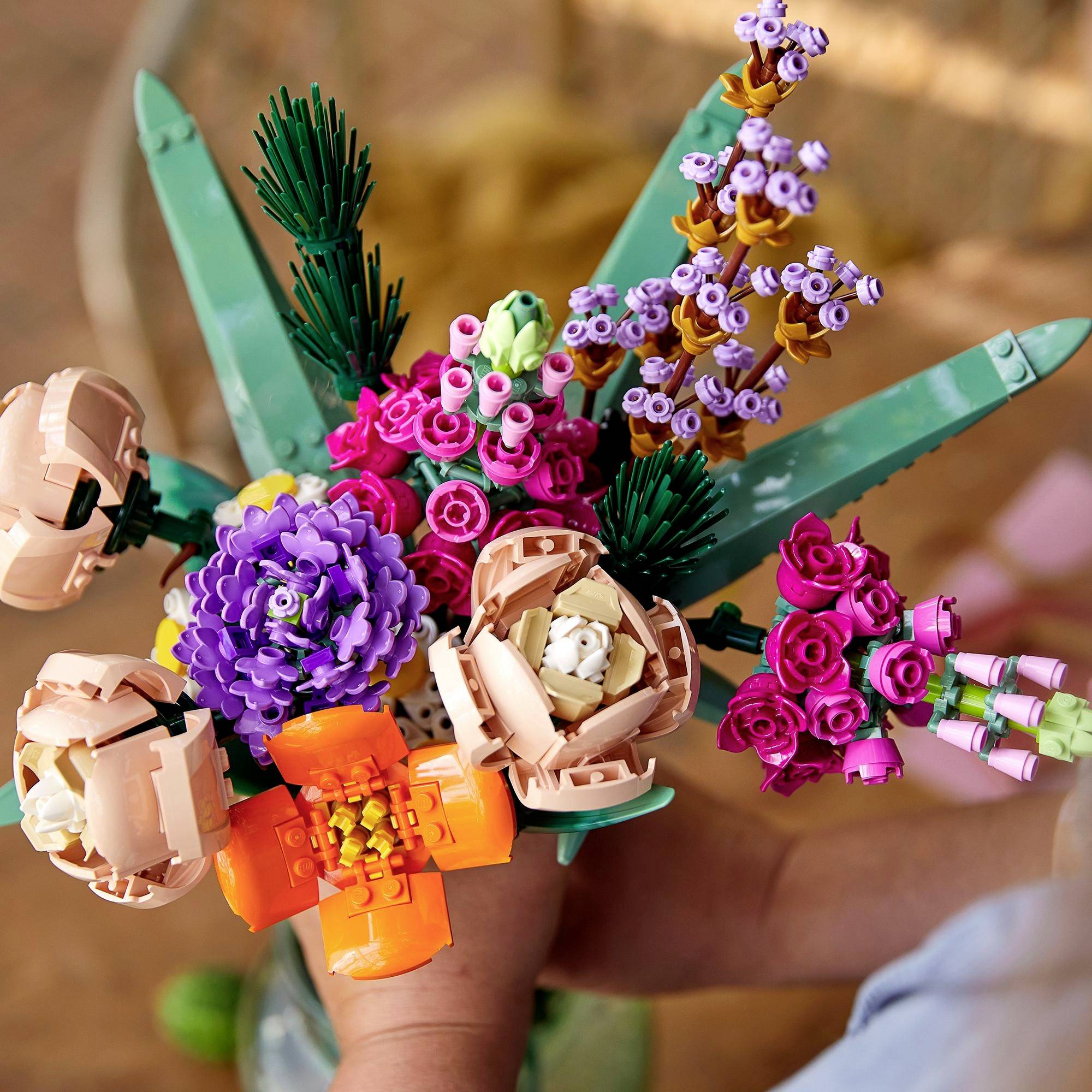 A person is holding a colourful bouquet of LEGO flowers with various colours and shapes, including purple, orange, and pink.