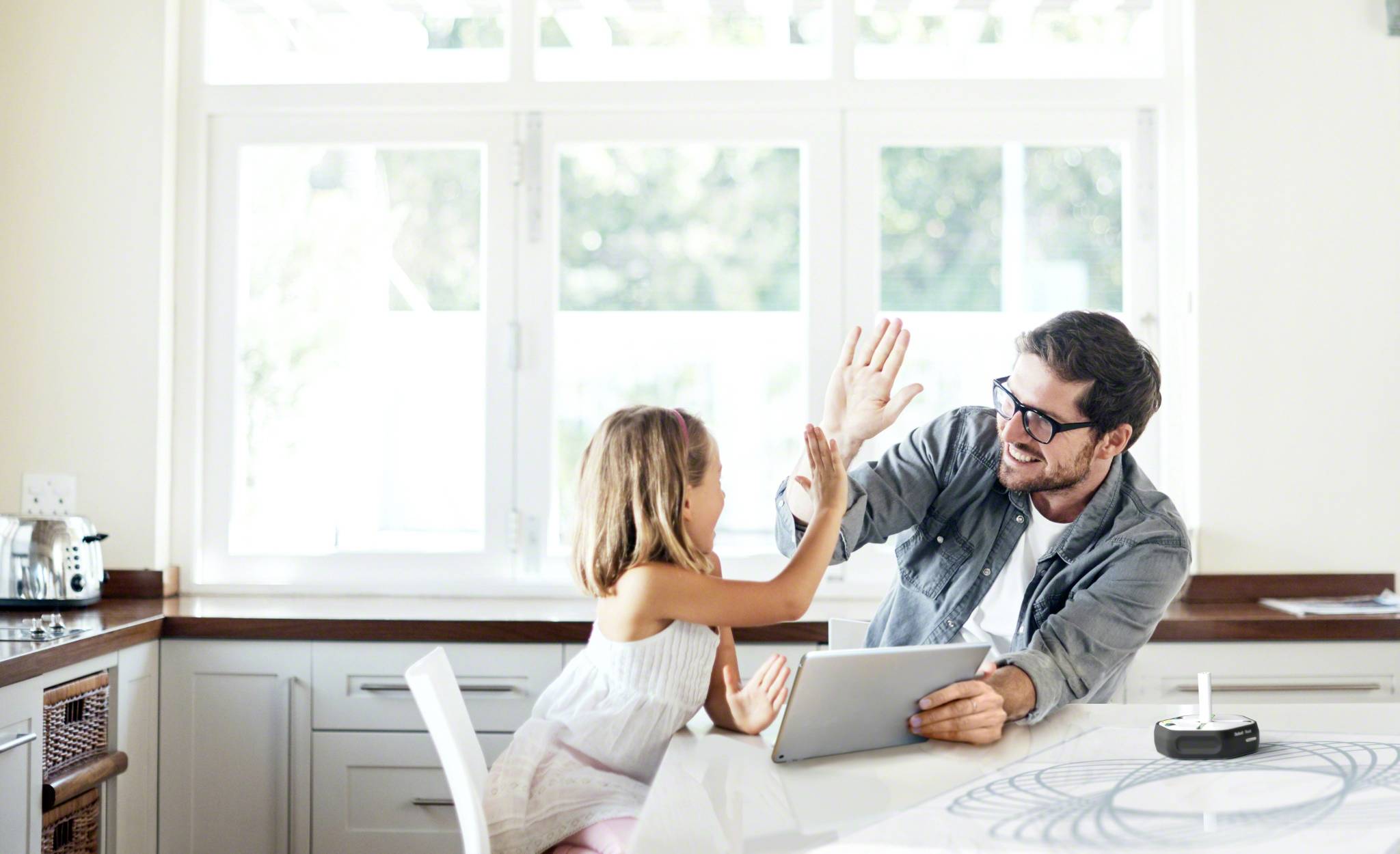 A man and a child are sitting in the kitchen, smiling at each other and giving a high-five. The man is holding a tablet. The atmosphere is cheerful.