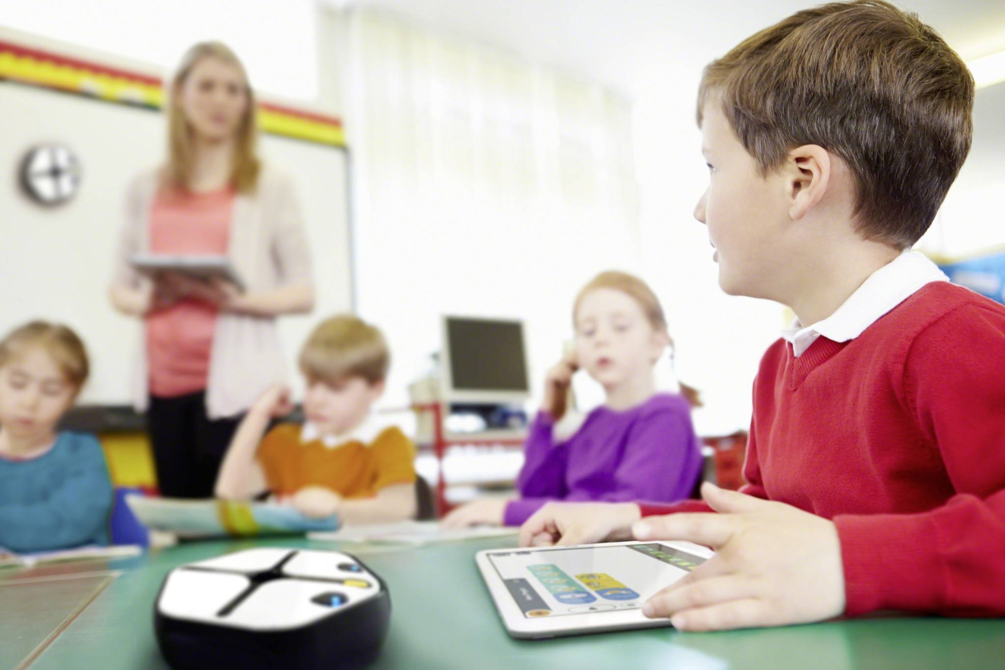 A boy in a red jumper is holding a tablet in a classroom. In the background, three children are seated and a teacher is standing with a tablet.