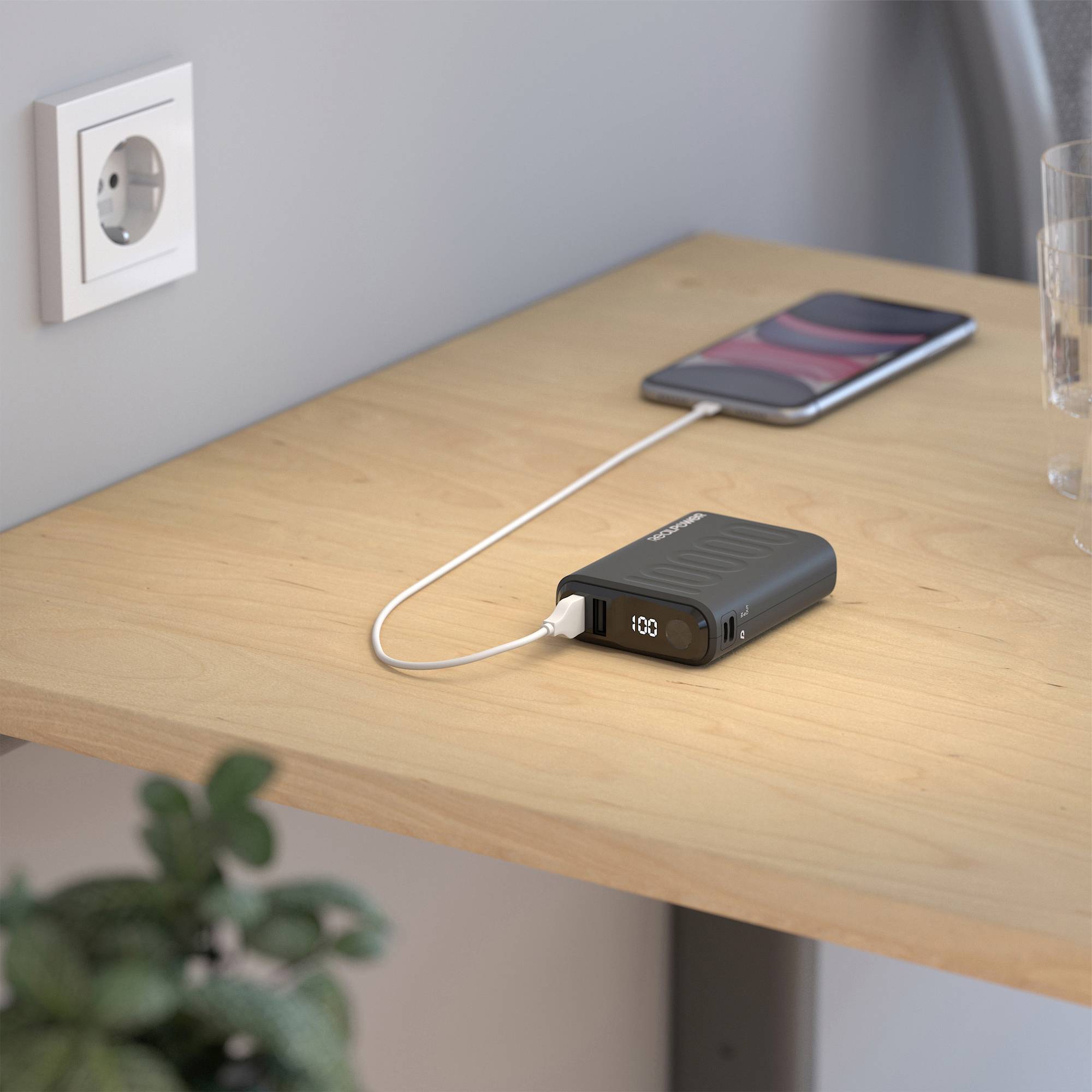 A mobile phone is being charged with a power bank on a wooden table. The power bank displays 100%. A plug socket is on the wall.