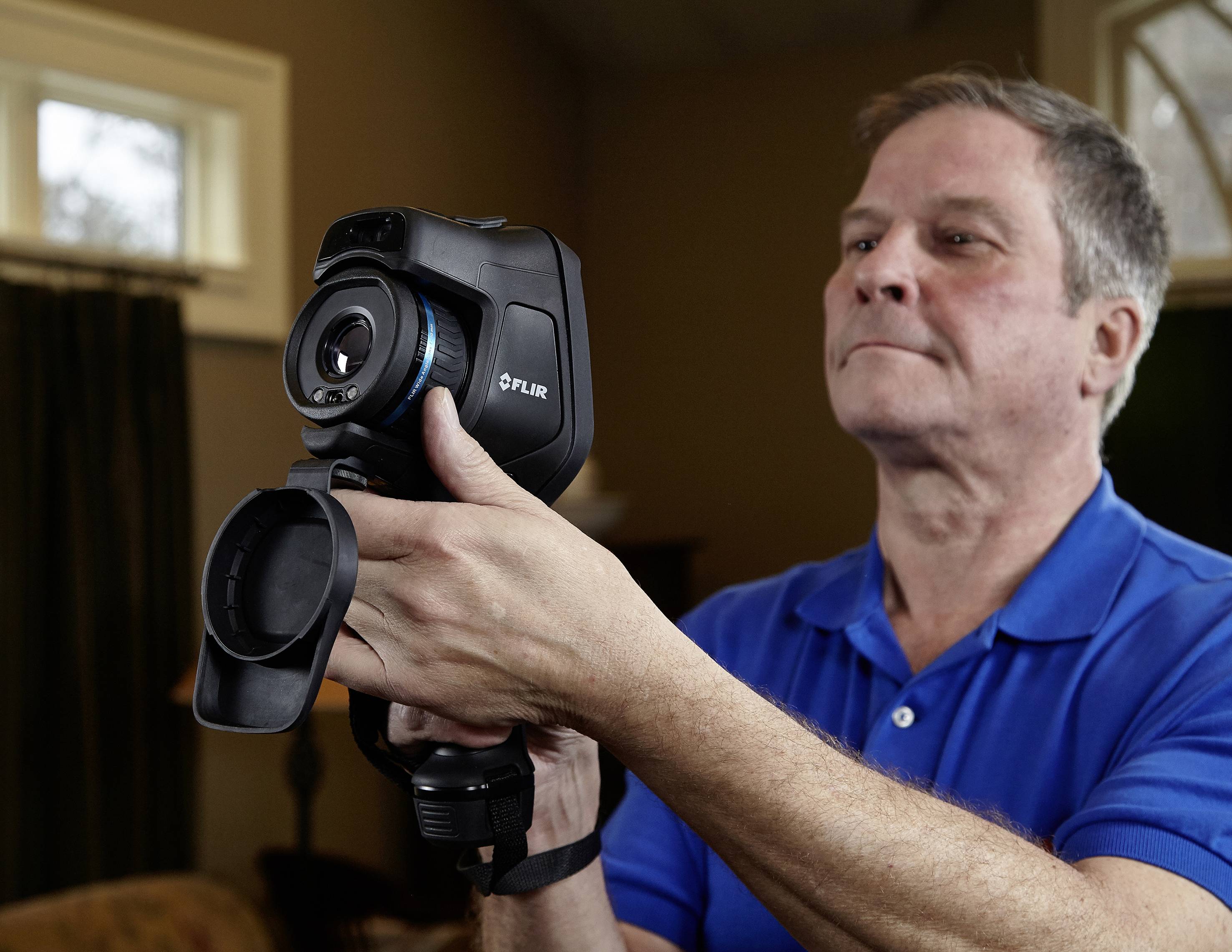 A man in a blue polo shirt is holding a thermal imaging camera inside a building. He is focused on capturing an image.