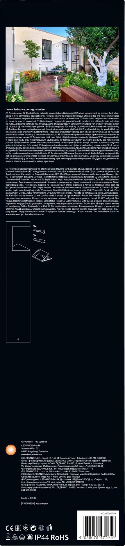 A white house with a wooden walkway leads to a door. To the right stands a large tree. The sky is blue and clear.