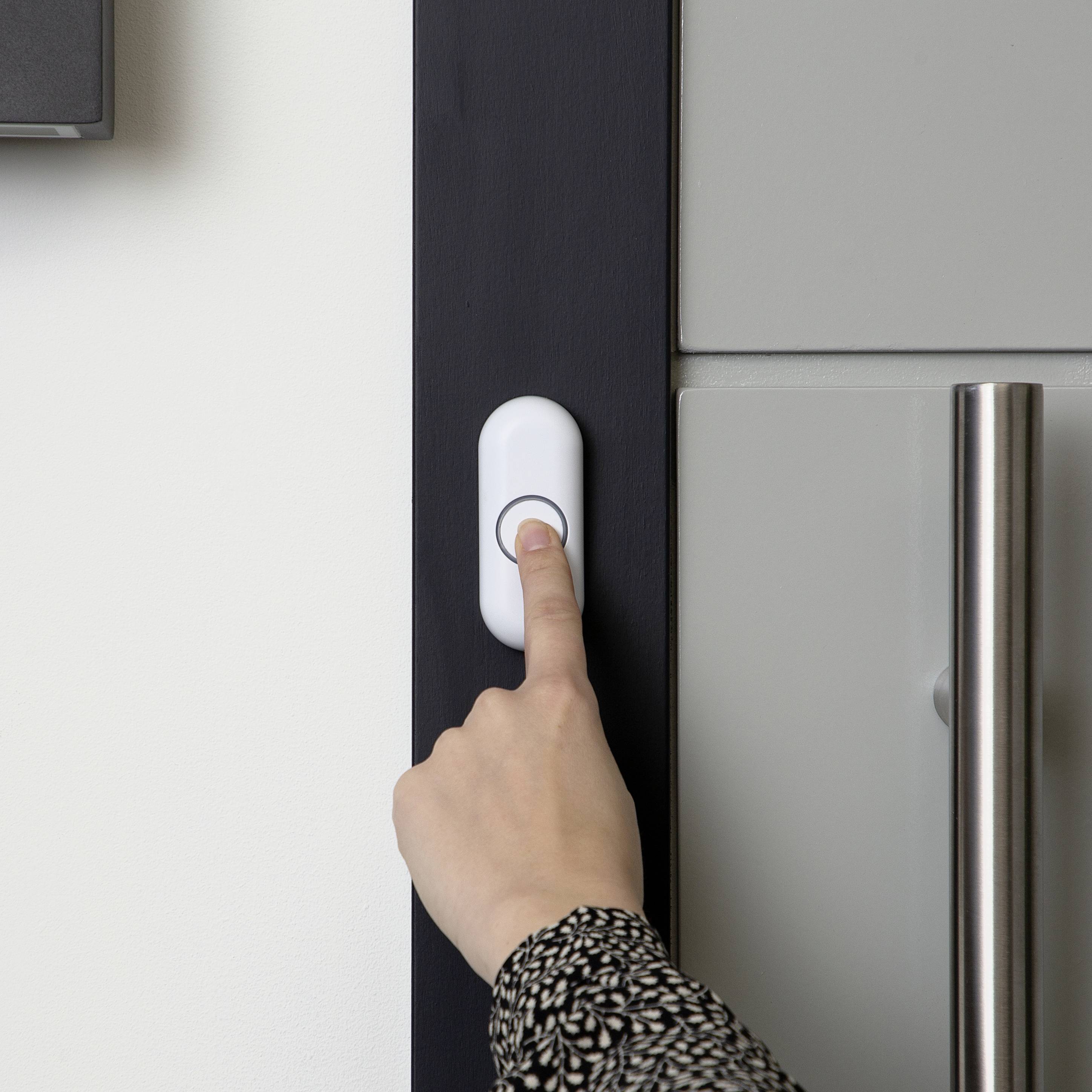 A hand is pressing a white doorbell on a black door frame. Next to the bell, a grey door with a metal handle is visible.