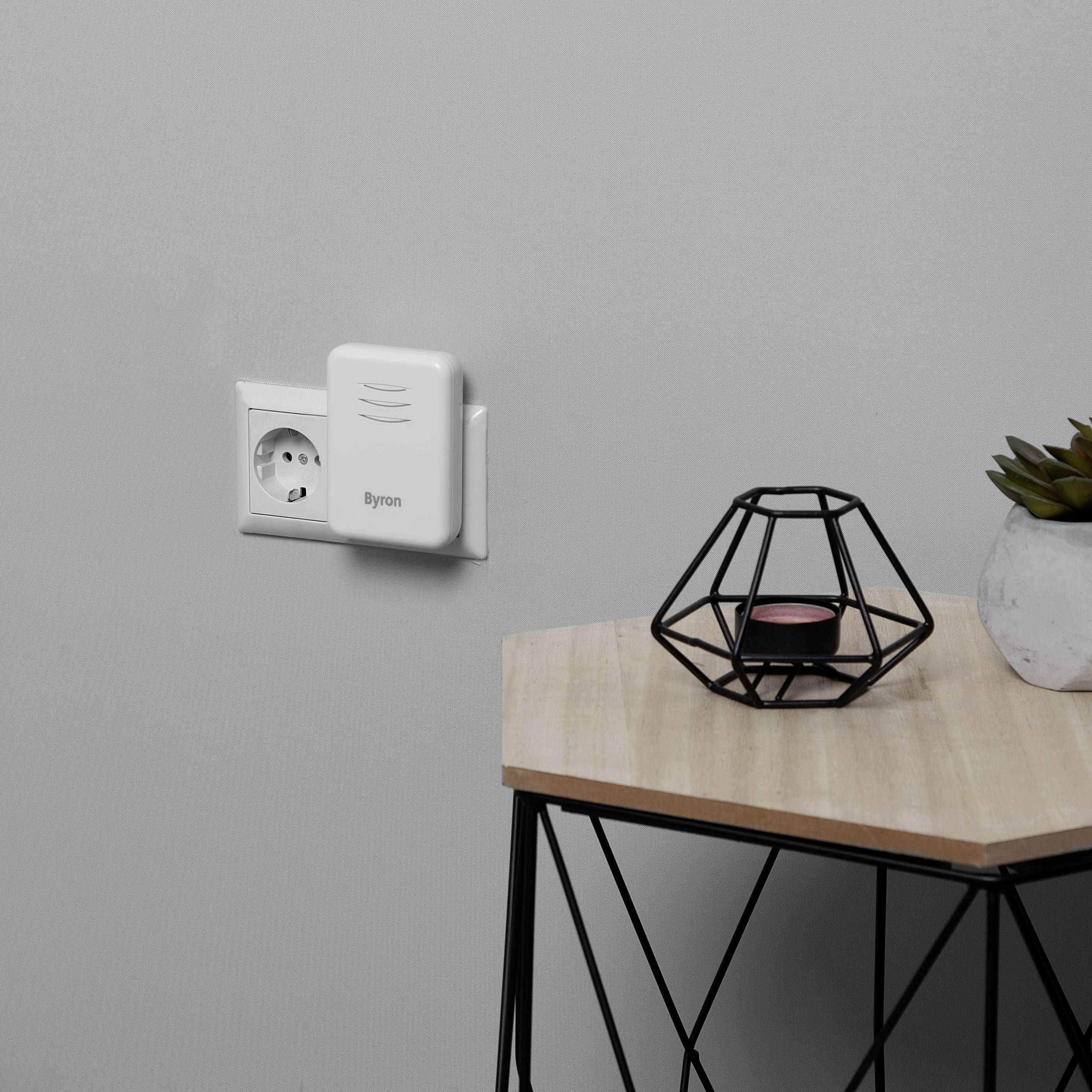 A white device is plugged into a wall socket. Next to it on a wooden table stands a decorative metal grill with a candle.