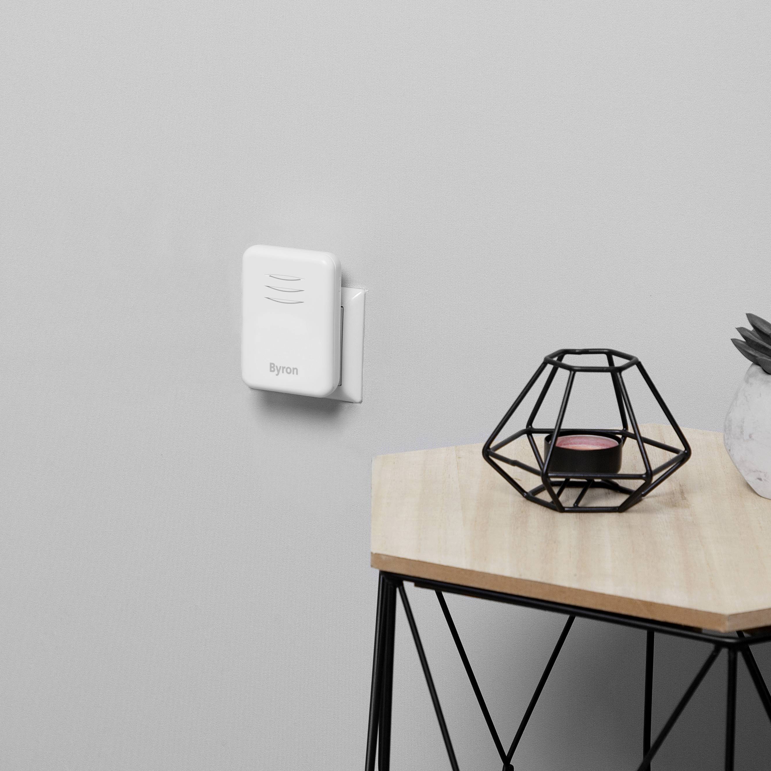 A white wireless doorbell receiver is mounted on a grey wall. Next to it, a decorative metal grid with a candle sits on a table.