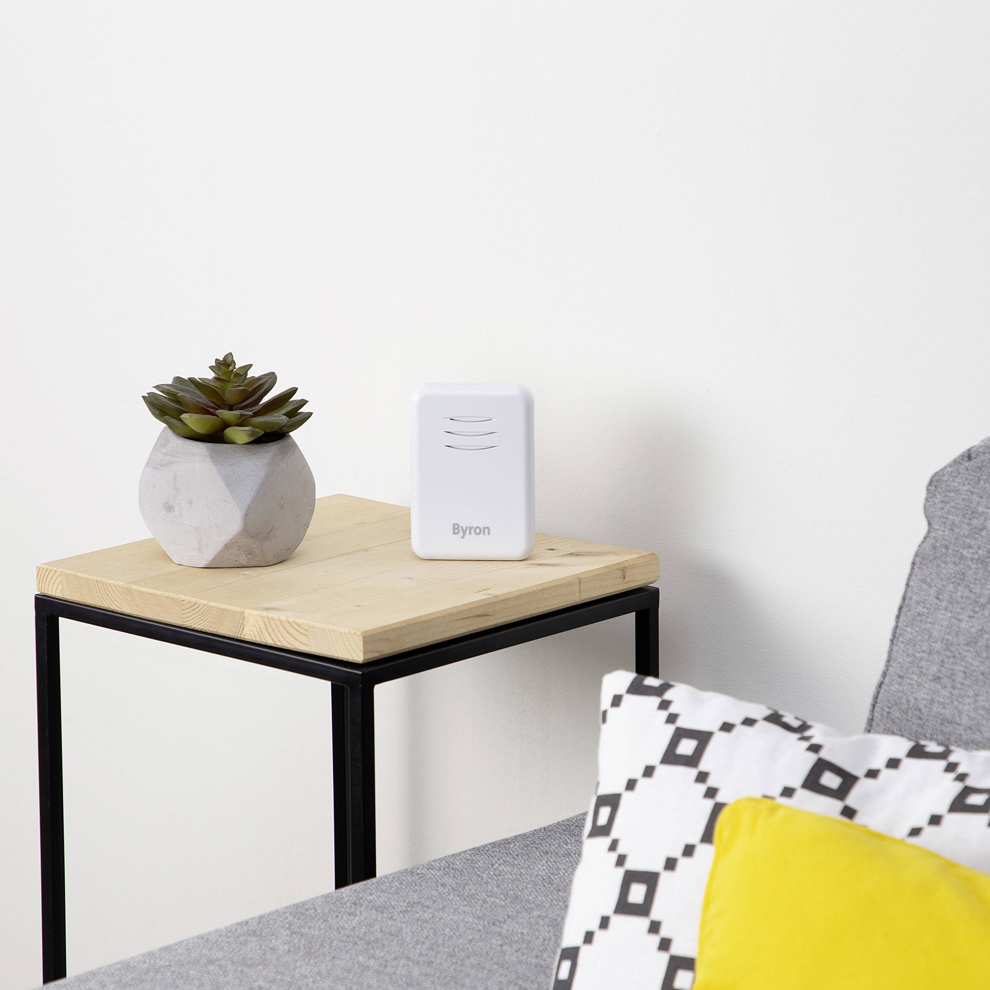 A square wooden table with a small white device and a potted plant stands beside a grey sofa with patterned cushions.