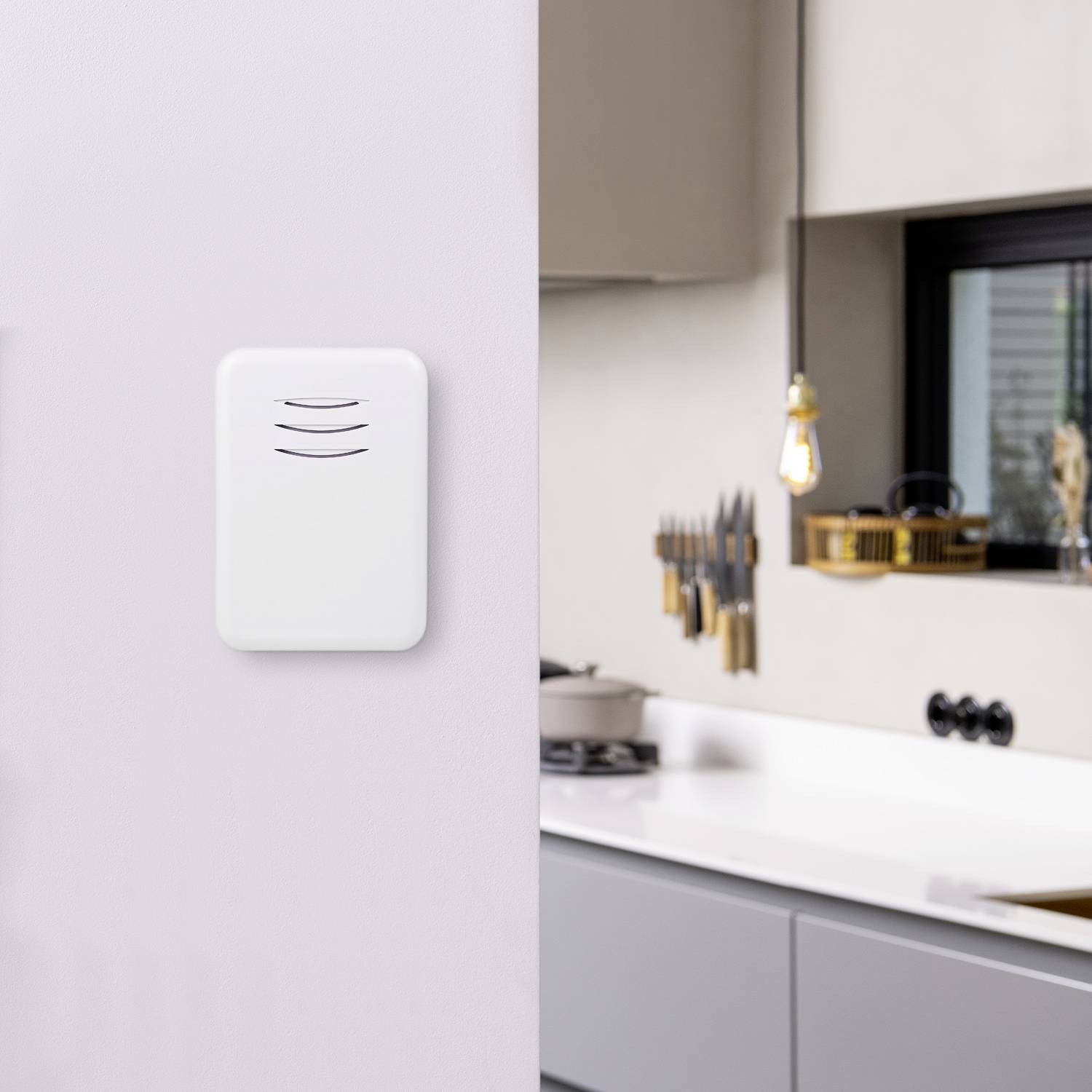 A white indoor air quality sensor mounted on a wall in a modern kitchen with minimalist design and subtle pendant lighting.