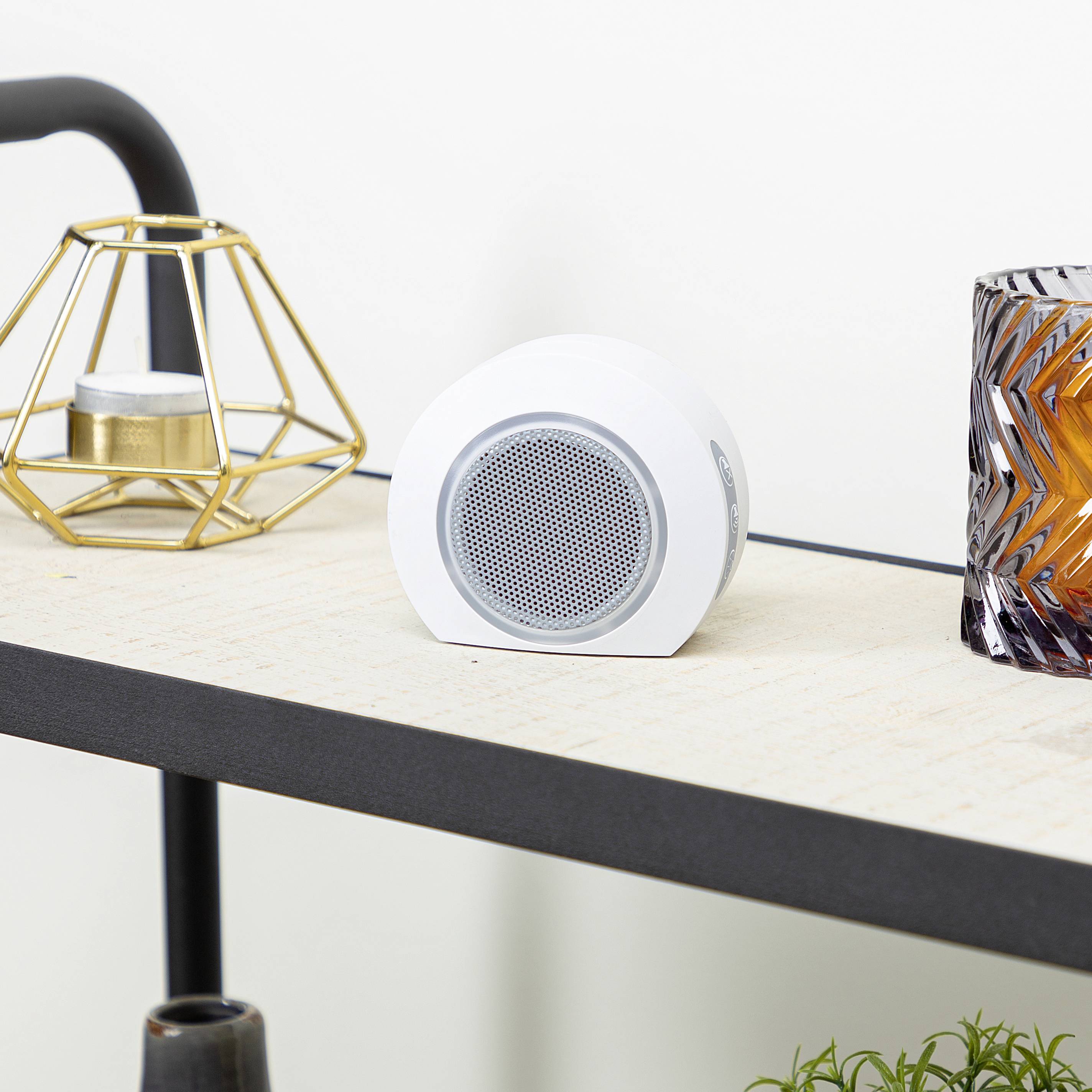 A white, round speaker sits on a shelf next to a geometric candleholder and a glass with zigzag-patterned designs.