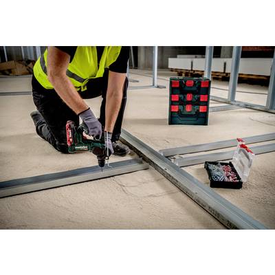 A tradesman is installing metal frames on a concrete floor using a drill. In the background, toolboxes with screws are visible.