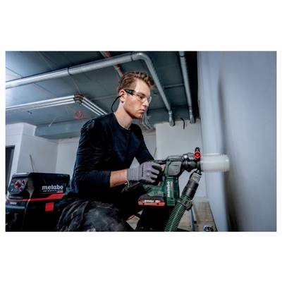 A person wearing protective goggles and gloves uses a Metabo power drill to work on a wall in a construction environment.