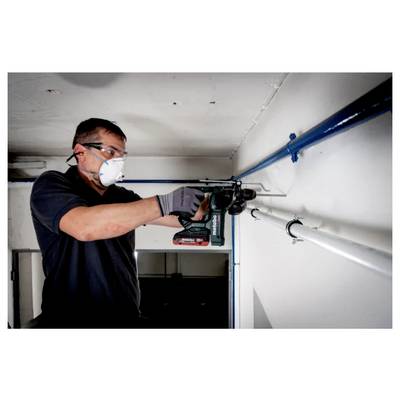 A person wearing safety goggles and a mask is using a power drill to bore a hole in a wall, focusing on their task in a small room.
