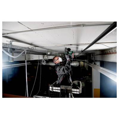 A person wearing a mask and safety gear stands on a platform installing or repairing a ceiling fixture in an industrial setting.
