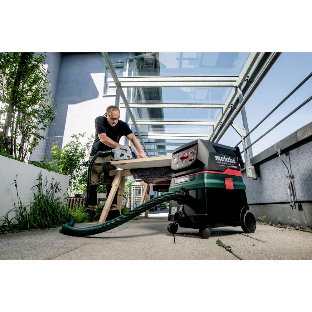 A person working outside using a power tool connected to a Metabo vacuum system on a patio. Glass roof and plants in the background.