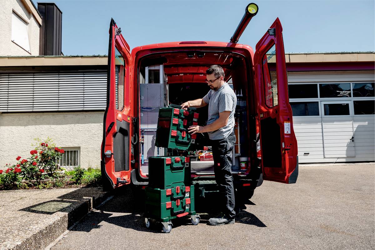 A man is unloading tools from a red delivery van. He is stacking green crates on a trolley in front of a garage.